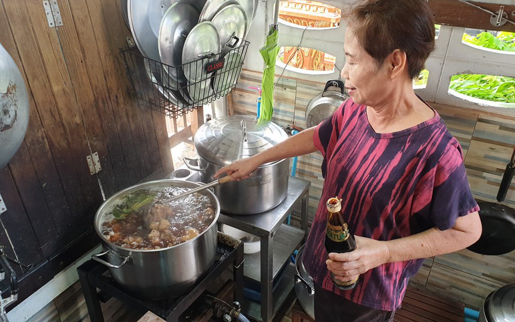 เจ้าของร้านมือหนึ่งกำลังใช้กระบวยตักซุปคนในหม้อขาหมูที่อยู่บนเตา ส่วนอีกมือหนึ่งถือขวดซอสปรุง